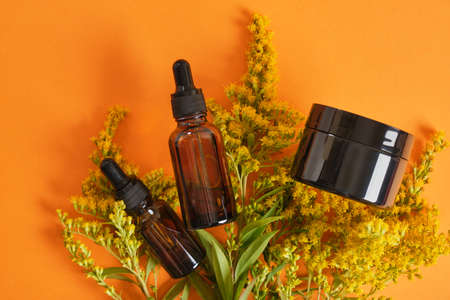 Black blank mock up cream jar and amber dropper glass bottles and yellow mimosa flower on orange background. Zero waste eco friendly spa product. Organic skin care cosmetic.の写真素材