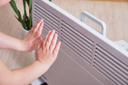 a small child warms his hands by the electric radiator in the room, the heating season is in the cold seasonの写真素材