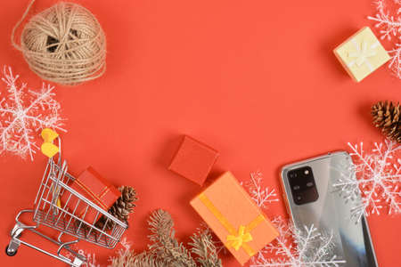 shopping cart and smartphone with three cameras, christmas decor on a red background, gifts boxes, snowflakes and phone, christmas and new year discounts and gifts conceptの写真素材