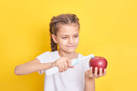 beautiful brown-haired teenage girl holding an apple and brushing her teeth with an electric toothbrush on a yellow background, children nutrition and oral hygiene conceptの写真素材