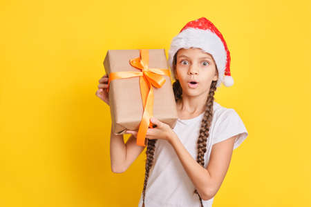 emotional cute teenager brown-hair girl in santa claus hat with a gift on a yellow background, christmas gifts concept copy spaceの写真素材