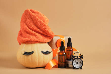 pumpkin with towel and false eyelashes, amber glass dropper bottles, gift box with ribbon and alarm clock on beige background, christmas or thanksgiving day promotion, body care conceptの写真素材