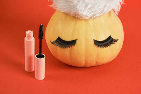 mascara in pink packaging mock up and pumpkin in santa hat and false eyelashes on red background winter cosmetic sale conceptの写真素材