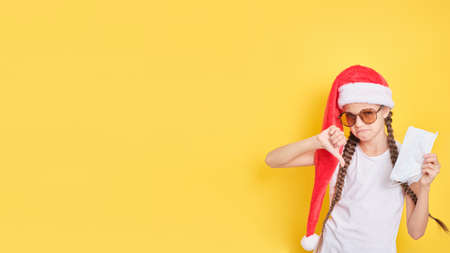 teenager girl in sunglasses and santa claus hat showing thumbs down gesture, yellow background, copy space, disgruntled girl wants to go on vacation for Christmas holidays, lockdown conceptの写真素材