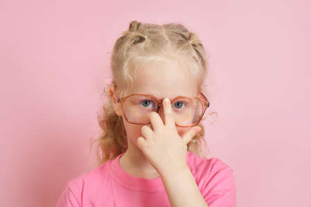 portrait of a cute caucasian preschooler girl in a pink T-shirt straightening glasses on her nose, children's ophthalmology concept, back to schoolの写真素材