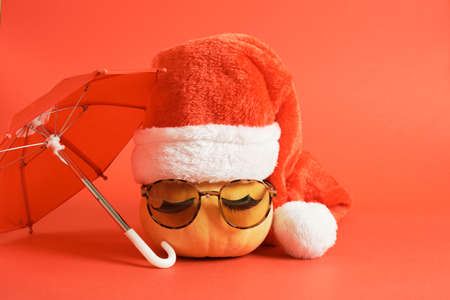 pumpkin with sunglasses and red santa claus hat under an umbrella on a red background, time for travel, christmas holidays in warm countries conceptの写真素材