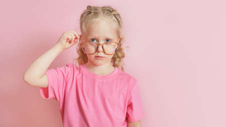 portrait of a cute caucasian preschooler girl in a pink T-shirt straightening glasses on her nose,...