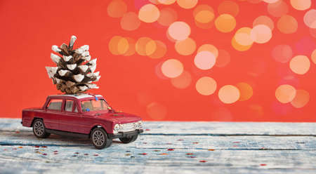 purple model of a retro car on a blue wooden surface with a pine cone on the roof, red background with bokeh, christmas background copy spaceの写真素材