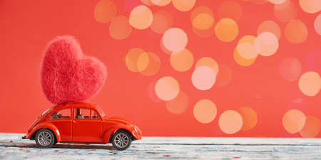 red retro car model on blue wooden surface with woolen heart on the roof, red background, christmas or valentine's day backgroundの写真素材