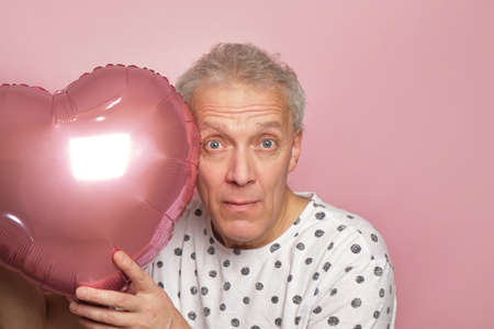 funny gray-haired elderly pensioner man holding pink heart-shaped balloon in his hands on pink...