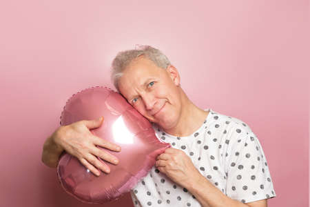 funny gray-haired elderly pensioner man holding pink heart-shaped balloon in his hands on pink background, valentines day conceptの写真素材