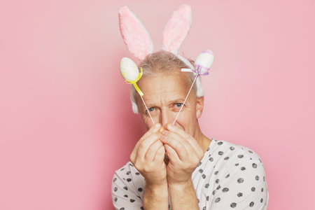funny gray-haired elderly pensioner man with bunny ears on his head and easter decorative eggs in his hands on a pink backgroundの写真素材