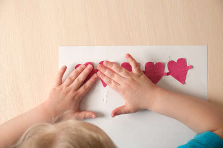 preschooler girl in blue t-shirt makes applique sitting at the table, hearts cut out of colored...