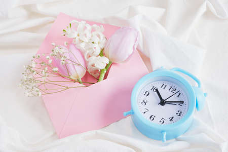 a bouquet of spring flowers in delicate shades, an alarm clock and a pink envelope on the bed, a romantic gift for a lover, March 8 or February 14 morning moodの写真素材