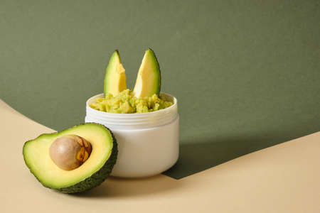 avocado half and white mockup blank jar with avocado puree on wooden stand, green background,...