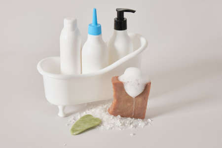 several different mock-up of white bottles with cosmetics in a miniature bathtub on a gray...
