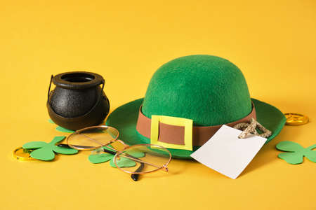 white blank tag, glasses, cauldron, gold coins, decorative green leprechaun felt hat from and leaves from foamiran on a yellow background, st. patricks day concept, copy spaceの写真素材