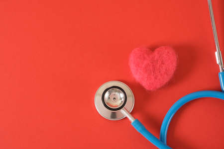 heart made of wool and a medical stethoscope on a bright red background copy space top view