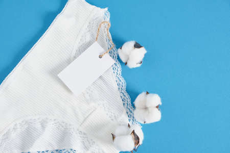 white cotton woman pants, blank paper label and cotton flowers on blue background copy space mock up