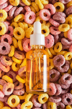 orange serum in a mock-up dropper glass bottle on the background of multi-colored breakfast cereals,...