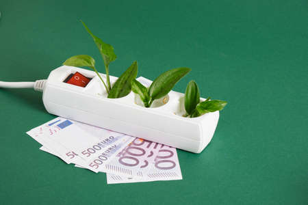 white surge protector with a red power button, money and green leaves of a plant on a green background, saving on electricity, electricity bill, eco-friendly energy resourcesの写真素材