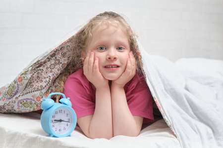 a cheerful preschooler girl in a pink t-shirt lies under the blanket and next to the alarm clock,...