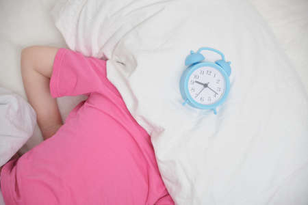 a preschooler girl in a pink t-shirt hides her head under a pillow, an alarm clock on the bed,...