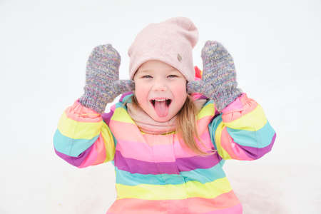 cheerful cute girl 5 years old plays in the snow and makes funny faces, shows tongue, child in winter in a bright striped suitの写真素材