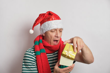 Positive senior man holding chhristmas gift box, wearing Santa hat on white background elderly man...