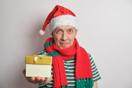 Positive senior man holding chhristmas gift box, wearing Santa hat on white background elderly man holding Christmas presentの写真素材