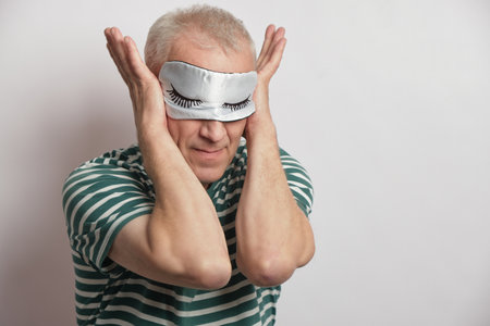 tired man in a sleep mask closes his ears on a gray background, insomnia of the elderly concept, annoying noise from neighbors does not let you sleepの写真素材