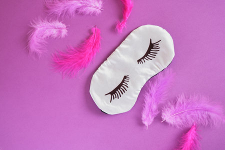 Top view silk sleep mask with painted eyelashes and bright pink and purple feathers, purple background copyspaceの写真素材