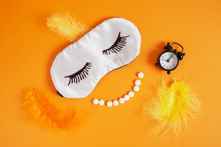 feathers, alarm clock, smile from pills and sleep mask with painted eyelashes on orange background top view insomnia conceptの写真素材