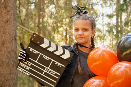 portrait of teenage girl in Halloween costume with balloons and movie clapperboard posing against natureの写真素材