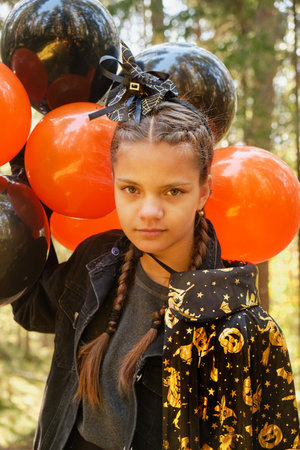 cute teenage girl with tanned skin in witch or wizard costume with bunch of orange and black balloons outside, halloween conceptの写真素材
