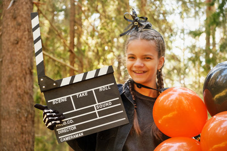 portrait of teenage girl in Halloween costume with bunch of balloons and movie clapperboard posing against background of treesの写真素材