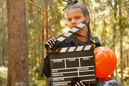 portrait of teenage girl in Halloween costume with bunch of balloons and movie clapperboard posing against background of treesの写真素材