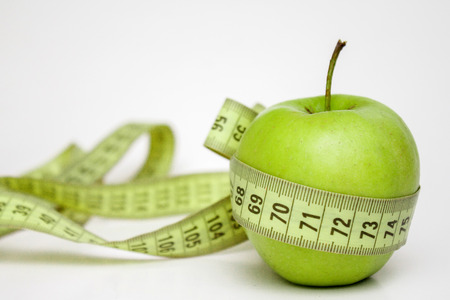 diet, healthy eating, food and weigh loss concept. Measuring tape wrapped around green apple on white backgroundの写真素材