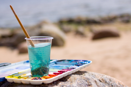 creation, drawing and freedom concept - Artistic equipment: paint brushes, tubes of paint, palette and paintings on rock in nature sea shore at sunny summer dayの写真素材