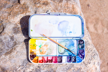 creation, drawing and freedom concept - Artistic equipment: paint brushes, palette and paintings on rock in nature at sunny summer dayの写真素材