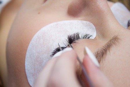 Eyelash Extension Procedure. Woman Eye with Long false Eyelashes. Close up macro shot of tweezers in...