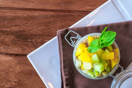 Healthy layered fruit dessert in glass jar. Kiwi, mango, pineapple, peach, chia seeds, mascarpone and whipped cream, mint and granola on brown wooden table. Top view. flat lay. Sweet menu conceptの写真素材