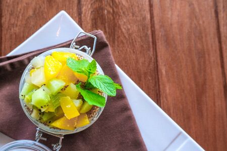 Healthy layered fruit dessert in glass jar. Kiwi, mango, pineapple, peach, chia seeds, mascarpone and whipped cream, mint and granola on brown wooden table. Top view. flat lay. Sweet menu conceptの写真素材