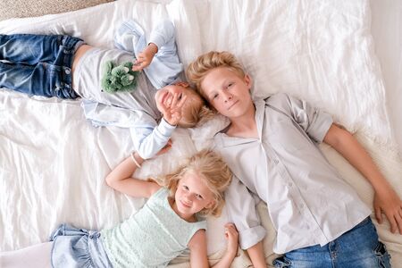 Three cheerful blonde kids are lying together on bed, looking at camera and smiling. Two boys and girl. Happy family, brothers and sister.の写真素材