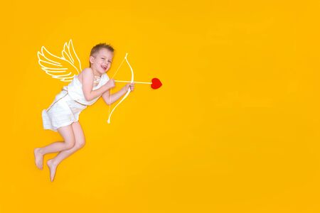 Happy smiling baby cupid in costume angel wings, bow and heart arrow isolated on yellow studio...