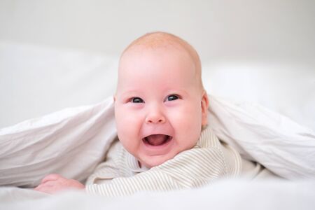 Adorable smiling baby boy under white blanket in bedroom. Newborn child relaxing in bed. Family morning at home. New born infant kids.の写真素材