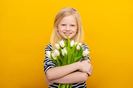 Delighted little girl with toothy smile, blonde hair, holds nice bouquet of white tulip flowers,...