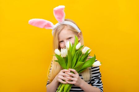 Girl in rabbit bunny ears on head on yellow studio background. Cheerful crazy happy child hidden behind tulips flower bouquet . Easter and spring.の写真素材