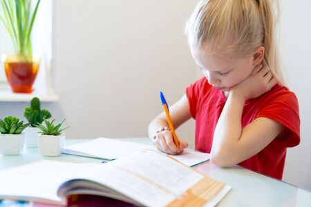 Serios blonde schoolgirl sitting at home and doing school homework, writing in notebook. Training...