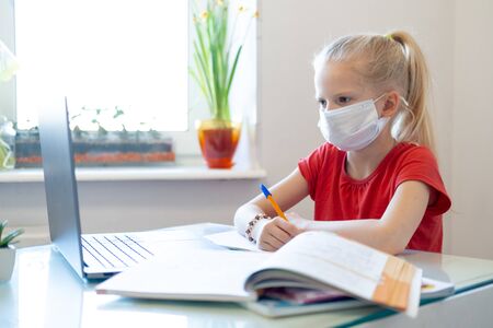 Distance learning online education. Sickness schoolgirl in medical mask studying at home, looking at laptop notebook and doing school homework. Training books and notebooks on tableの写真素材